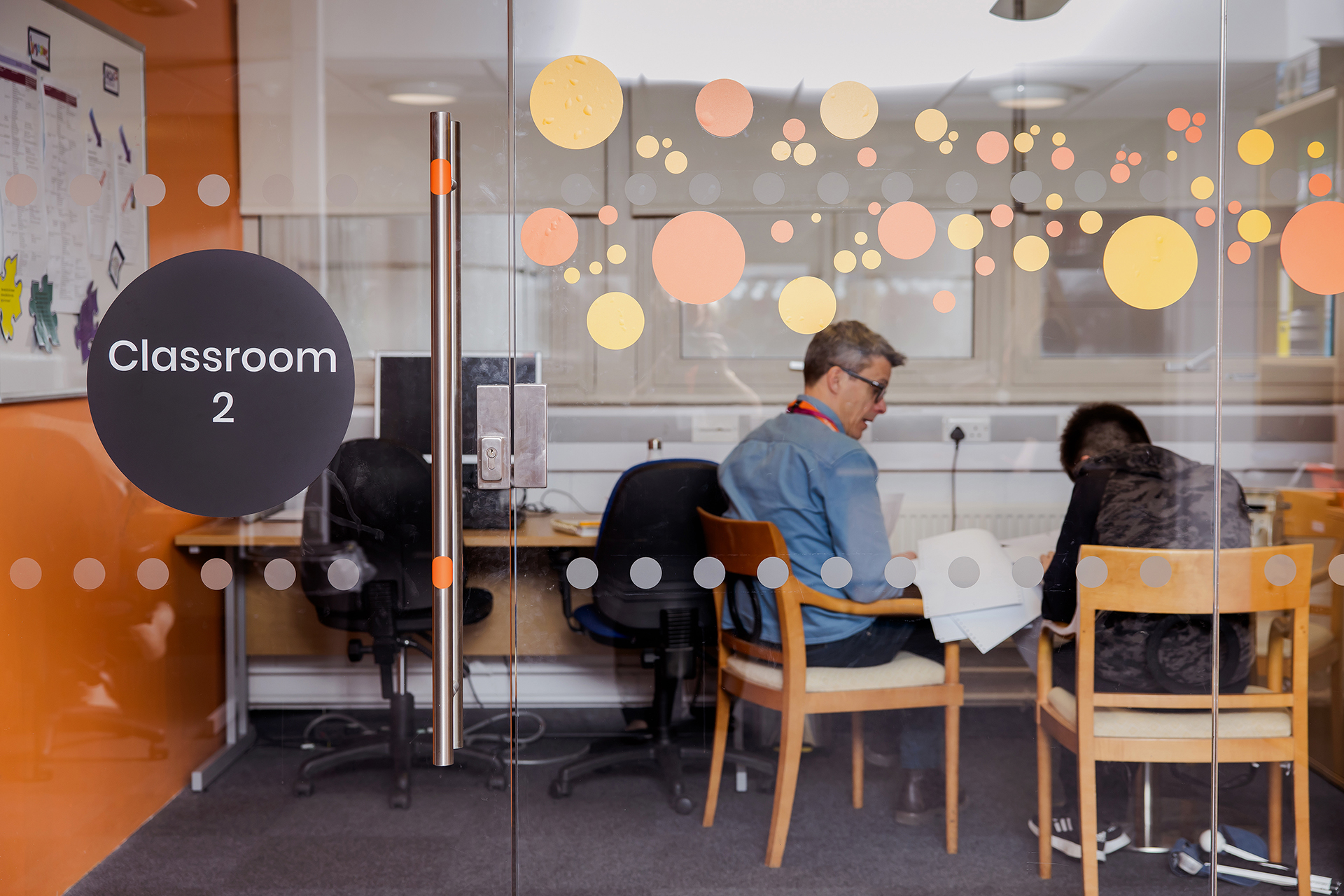 A male student and male tutor sat in a classroom with an glass wall.