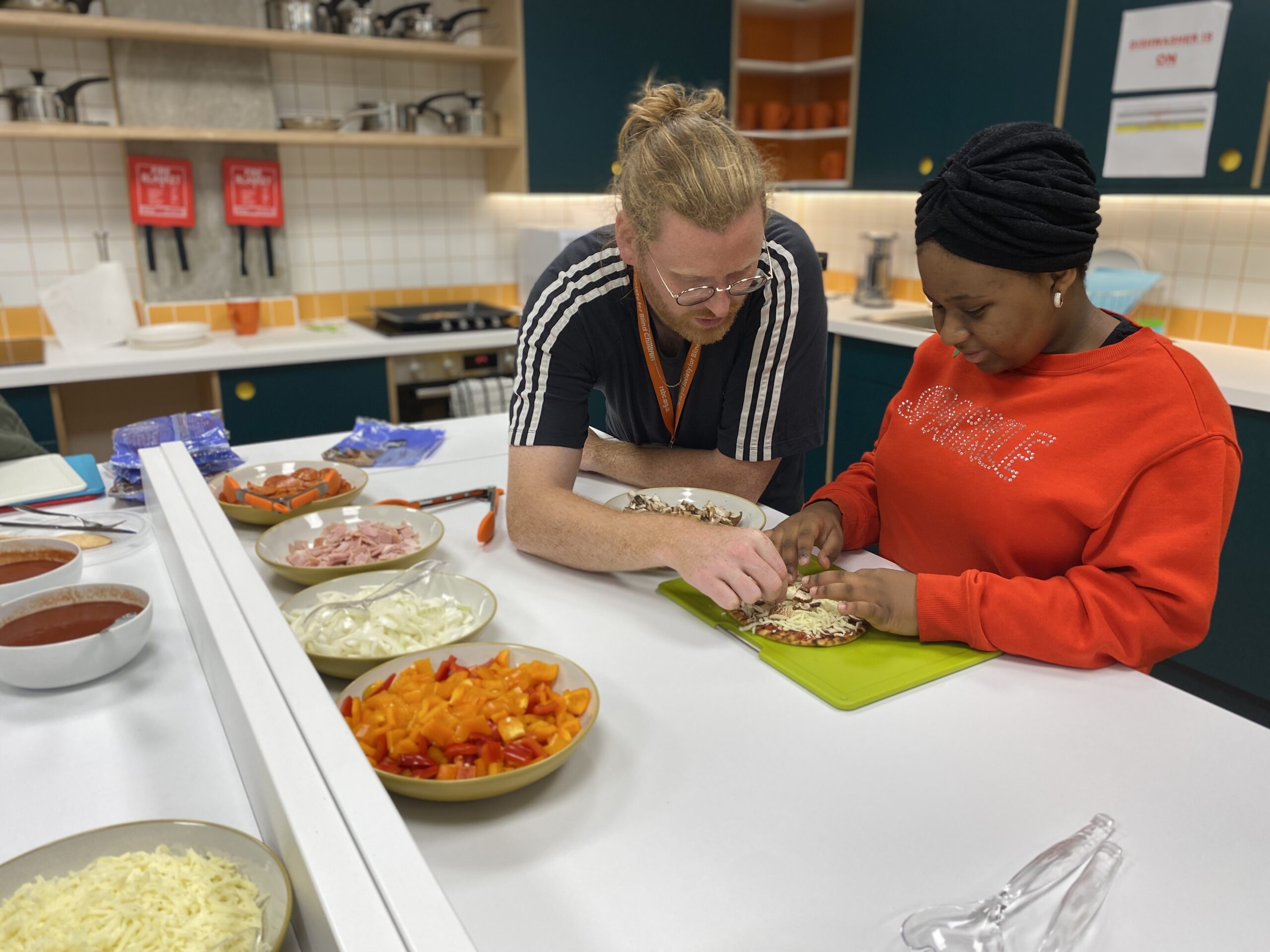 Two people in the RSBC kitchen making a pizza. On the left is Owain, a light-skinned man with glasses and long ginger hair tied up in a bun. He's wearing a navy t-shirt with an orange RSBC lanyard. On the right is a dark-skinned person, wearing a black velvet headwrap and a red jumper with 'sparkle' written across it in sequins.