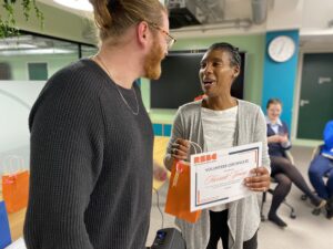 On the left is a light-skinned man with ginger hair, tied up in a top-knot. Next to him is a dark-skinned woman with short black hair. They are talking to each other, smiling. The woman is holding a certificate that read 'Volunteer Certificate presented to Harriett Young'.