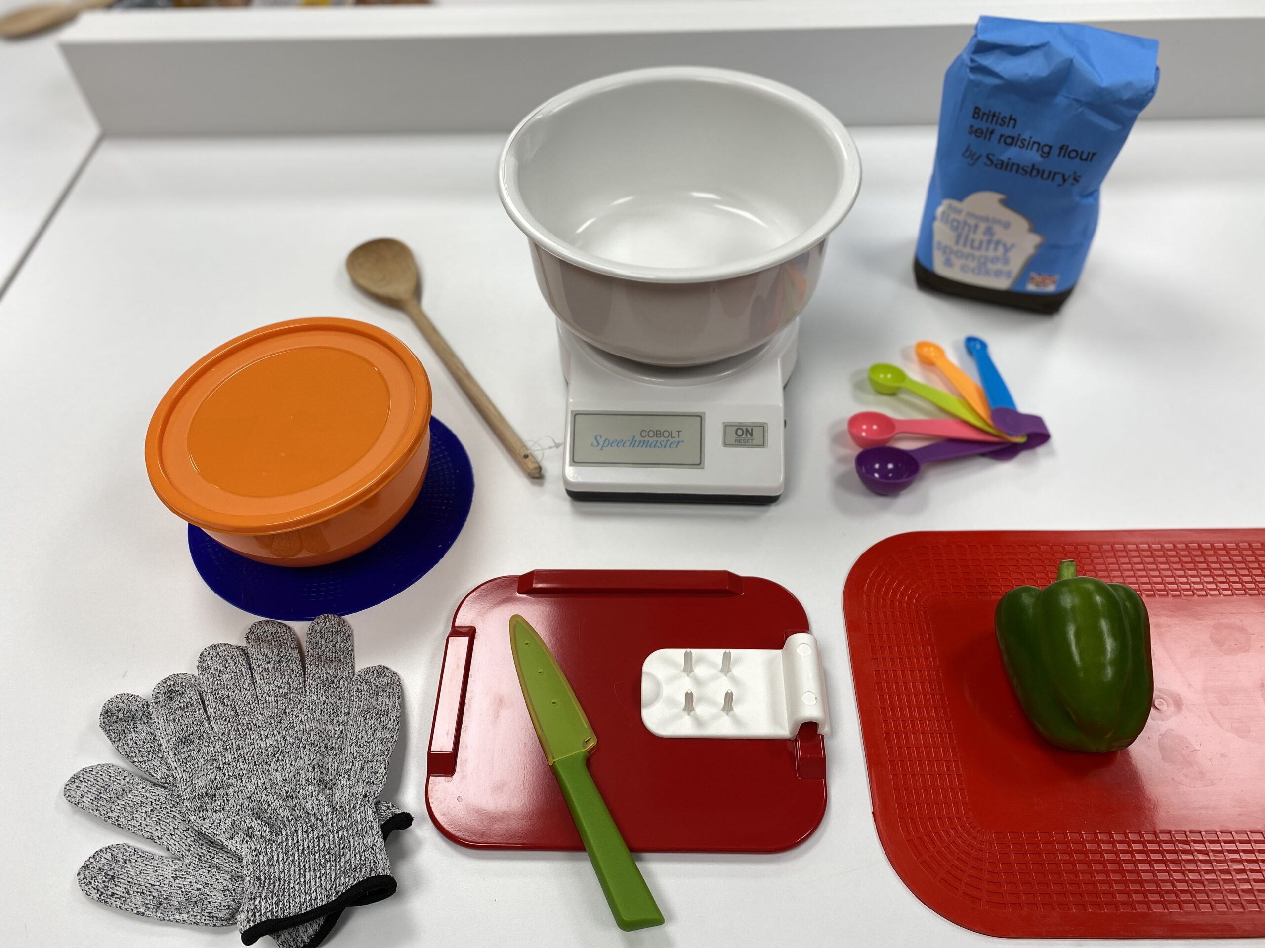 A range of kitchen equipment laid out on the counter. From top left to bottom right: a wooden spoon, a mixing bowl and kitchen scale, a bag of flour, an orange Tupperware tub, a red chopping board with a green kitchen knife, a red chopping board with a green pepper and a pair of safety gloves.