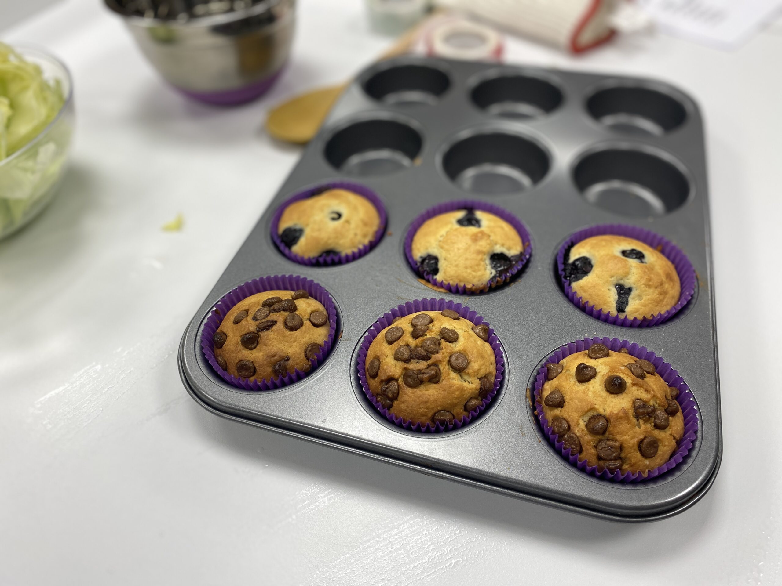 A metal muffin tray filled with delicious looking blueberry muffins and chocolate chip muffins. In the background there is a messy kitchen.