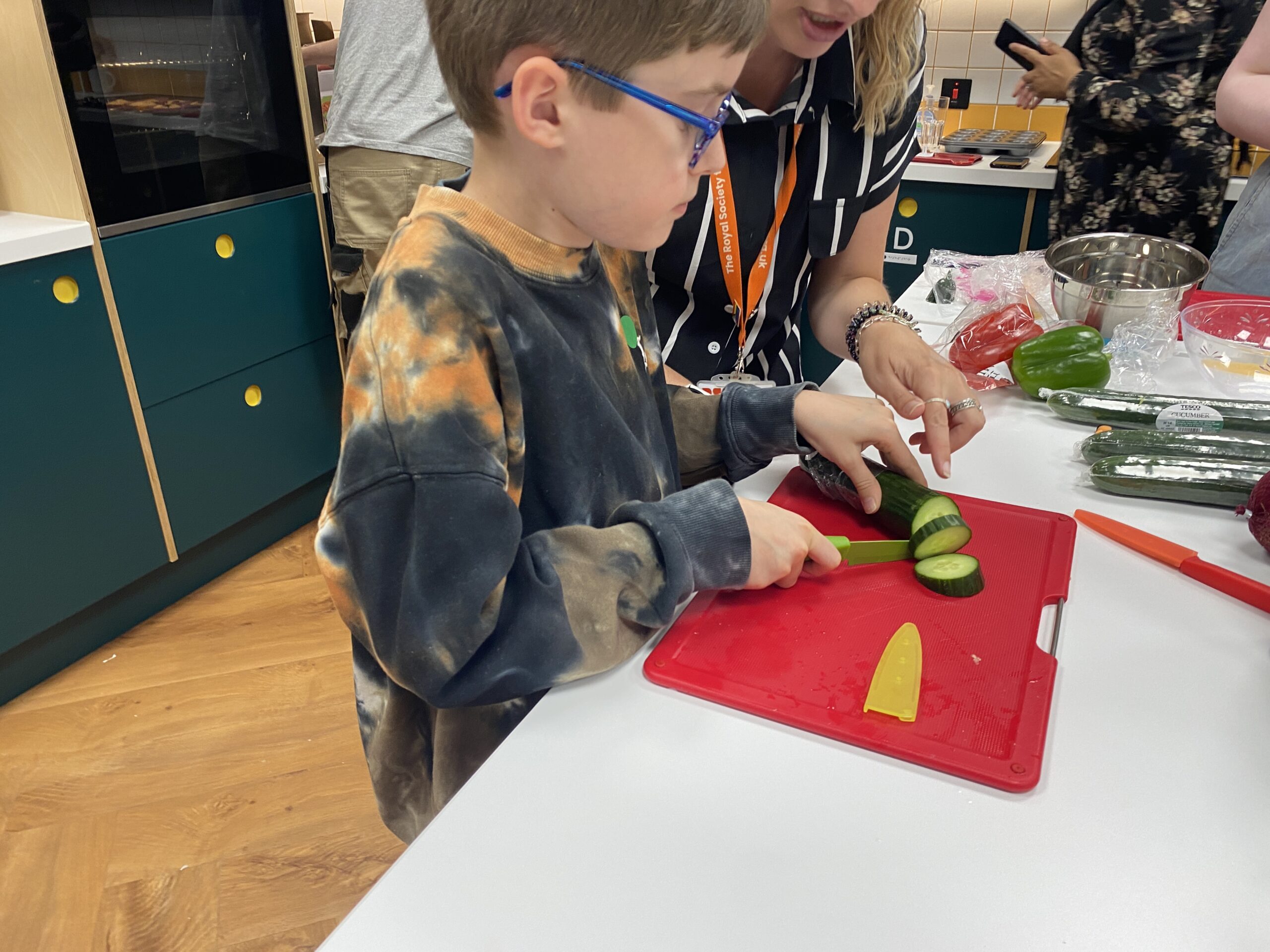 A light-skinned young boy with short brown hair and blue glasses, using a bright green cutting knife to cut cucumber on a red cutting tray in the RSBC kitchen.