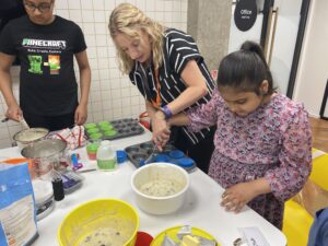 A lady in a kitchen holding onto and guiding the arm of a young medium-skinned girl