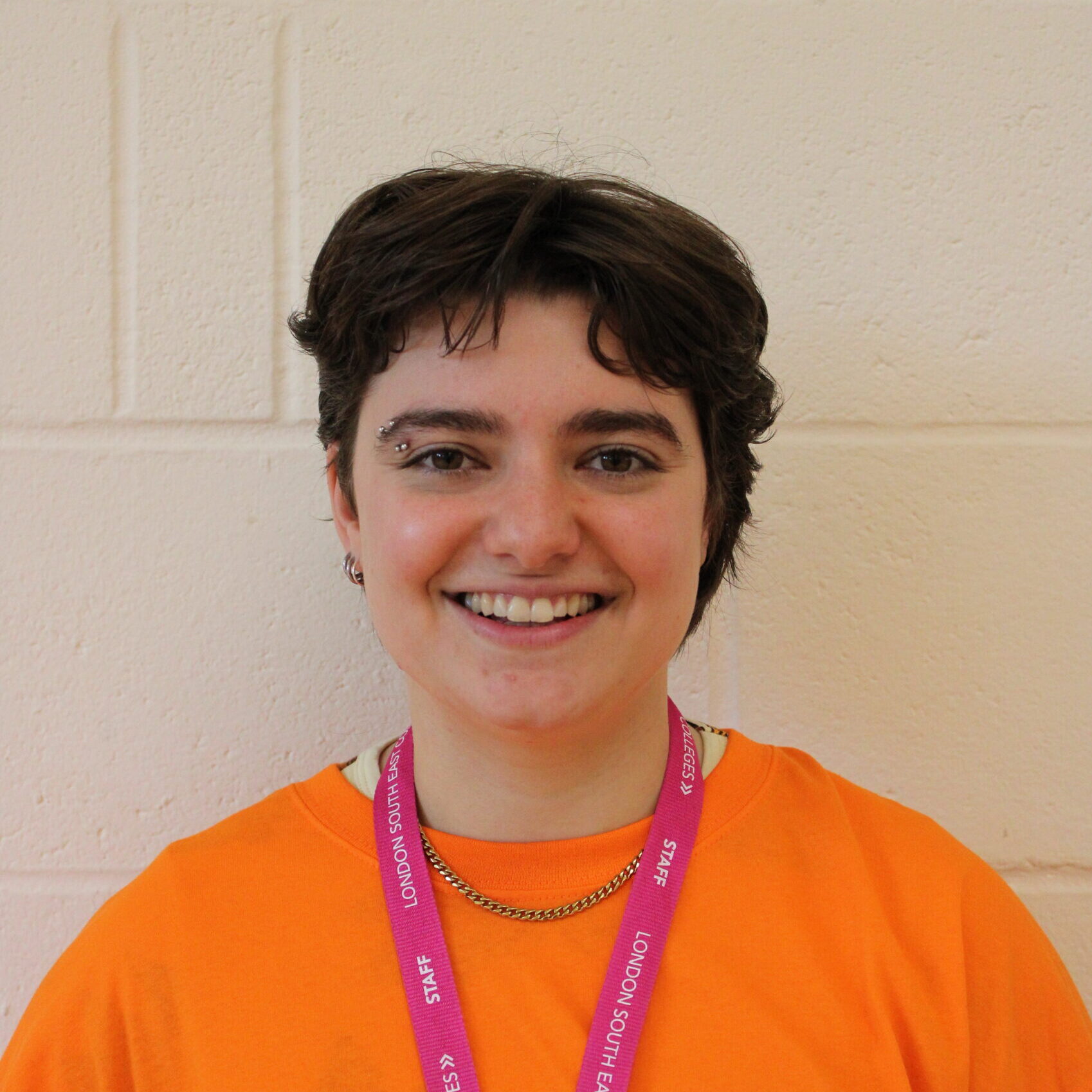 A headshot of Atrix, a light-skinned person with short brown hair and dark eyes. They are wearing an orange T shirt with a pink lanyard, have a silver eyebrow piercing over their right eye and are smiling cheerily at the camera.