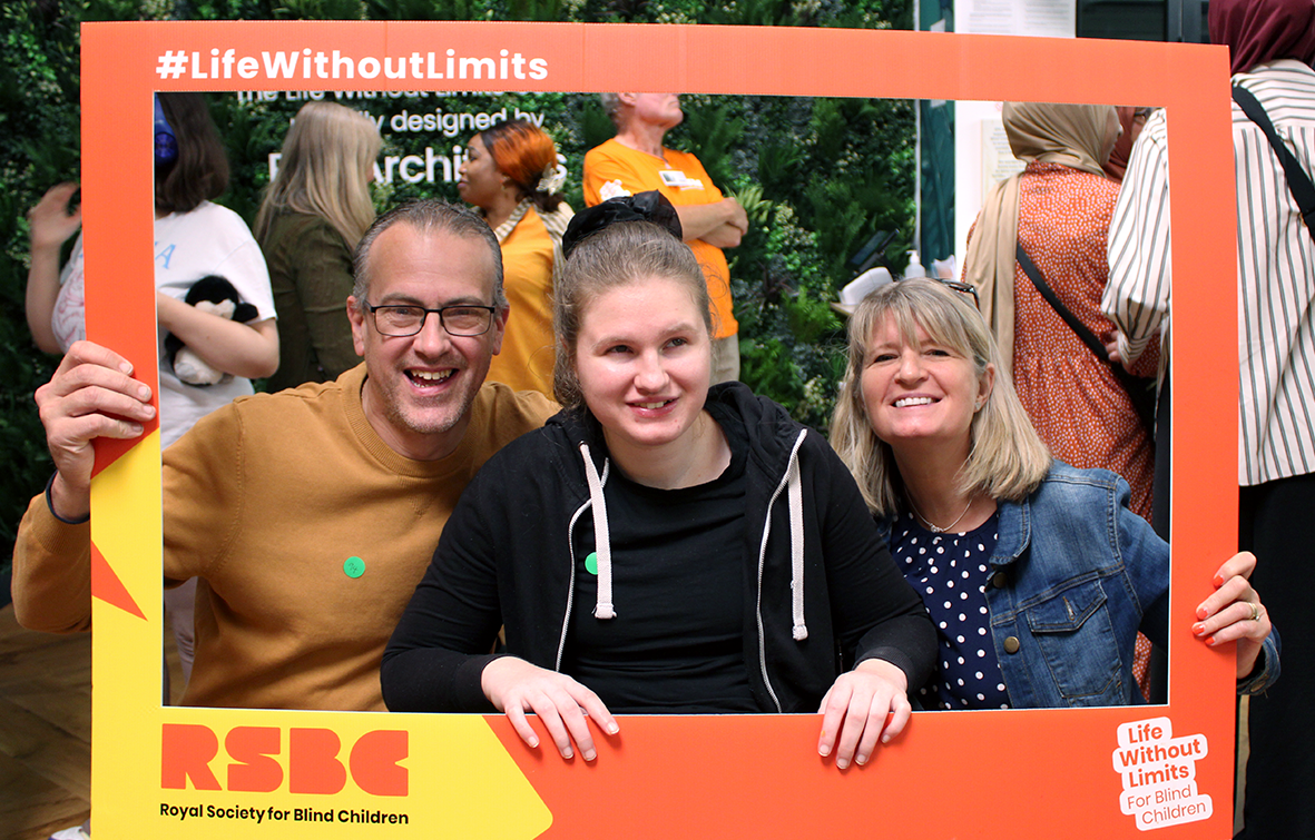 A family of three huddled together in a novelty selfie-frame, posing for a picture. From left to right: a light-skinned man with short grey hair and glasses, a light-skinned young woman with long light-brown hair held up in a ponytail, a light-skinned woman with medium length blonde hair. The people are all smilingly warmly at the camera.