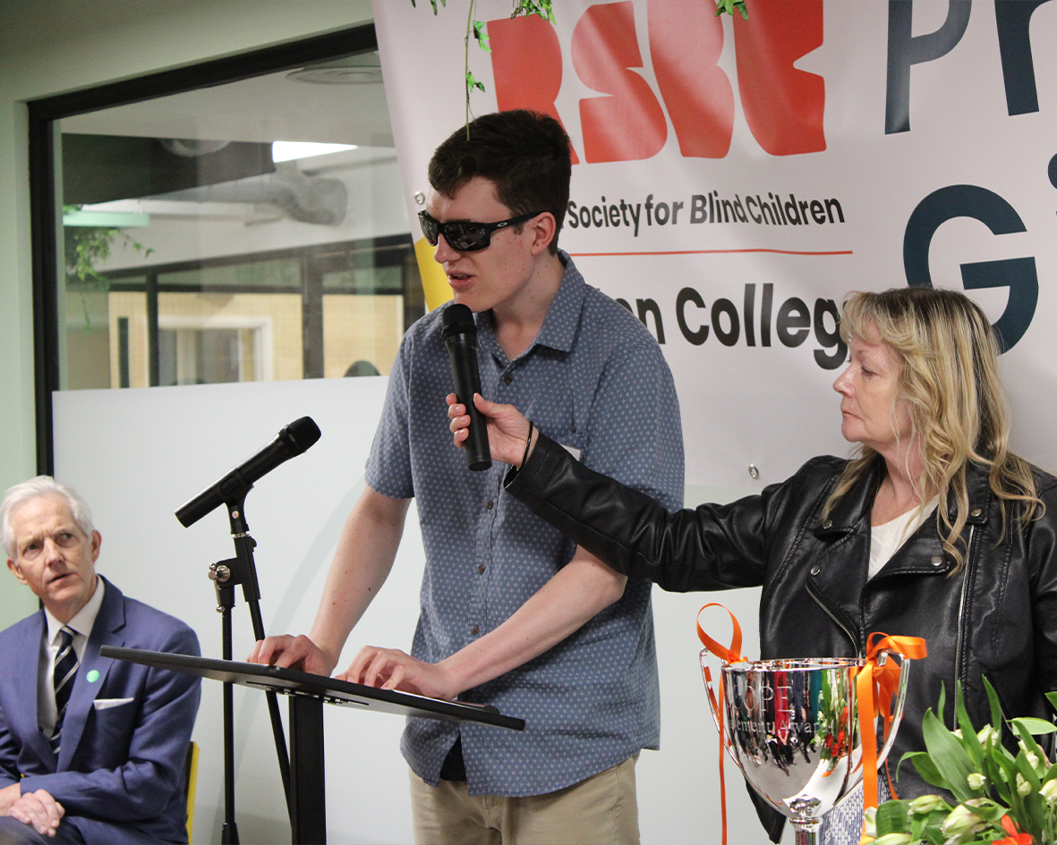 A young man with short brown hair and dark lensed-glasses. He is standing behind a podium. A woman with medium-long blonde hair, stands beside him holding a microphone to his face while he speaks. 