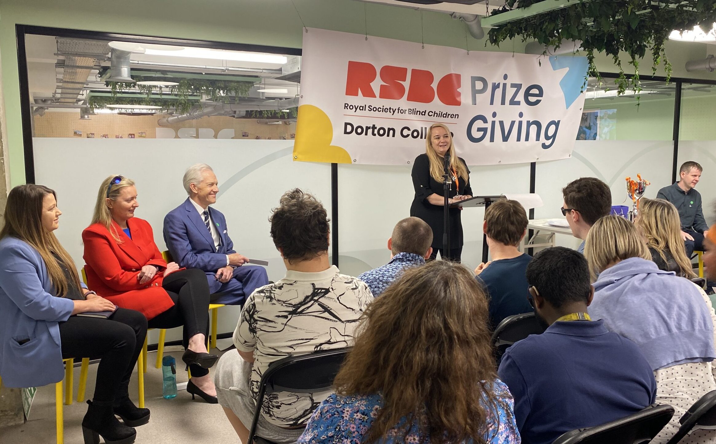 A light-skinned woman with straight long blonde hair, standing behind a podium, speaks into a microphone. To the left of her, sit a row of the guests. In front of here is an audience of people. 