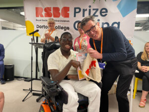 A student receiving a certificate from a teacher at the RSBC Dorton College Prize Giving ceremony 2024. The student, on the left, is a young dark-skinned person with short black hair. They are using a black motorised wheel-chair and are wearing a cream t-shirt with white jeans. The teacher, on the right, is a light-skinned person with short grey hair and glasses. They are tall but henched over to reach eyelevel with the student in the wheelchair. The two are smiling at the camera, whilst jointly holding a certificate.