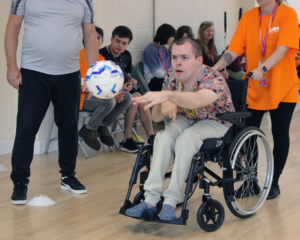 Action shot of Frankie, a Dorton College student, throwing a ball at a DC sports event. Frankie, a light-skinned young man with short brown hair, is sat leaning forward in his wheelchair with an intense look of concentration on his face.