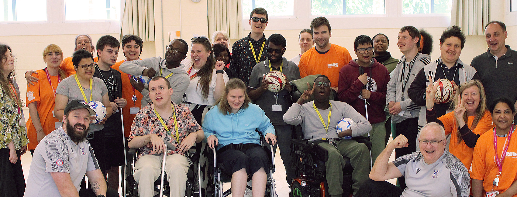 A group photo of the Dorton College students and staff at a sports activity day with Crystal Palace. There are approx. 30 people huddled into the shot. In the front row there are three students in wheelchairs, with the rest fo the group standing behind them. Everyone is smiling and cheering.