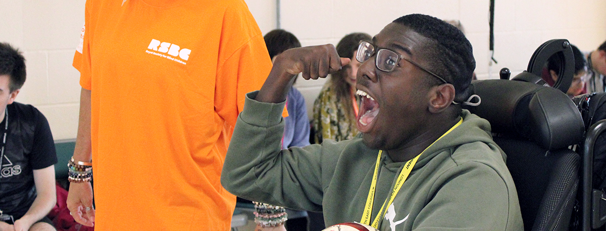 A dark-skinned young man in a wheelchair cheering victoriously as he celebrates an epic goal. With one arm, he holds a football and with the other he raises his fist in the air. He has very short black hair and glasses.