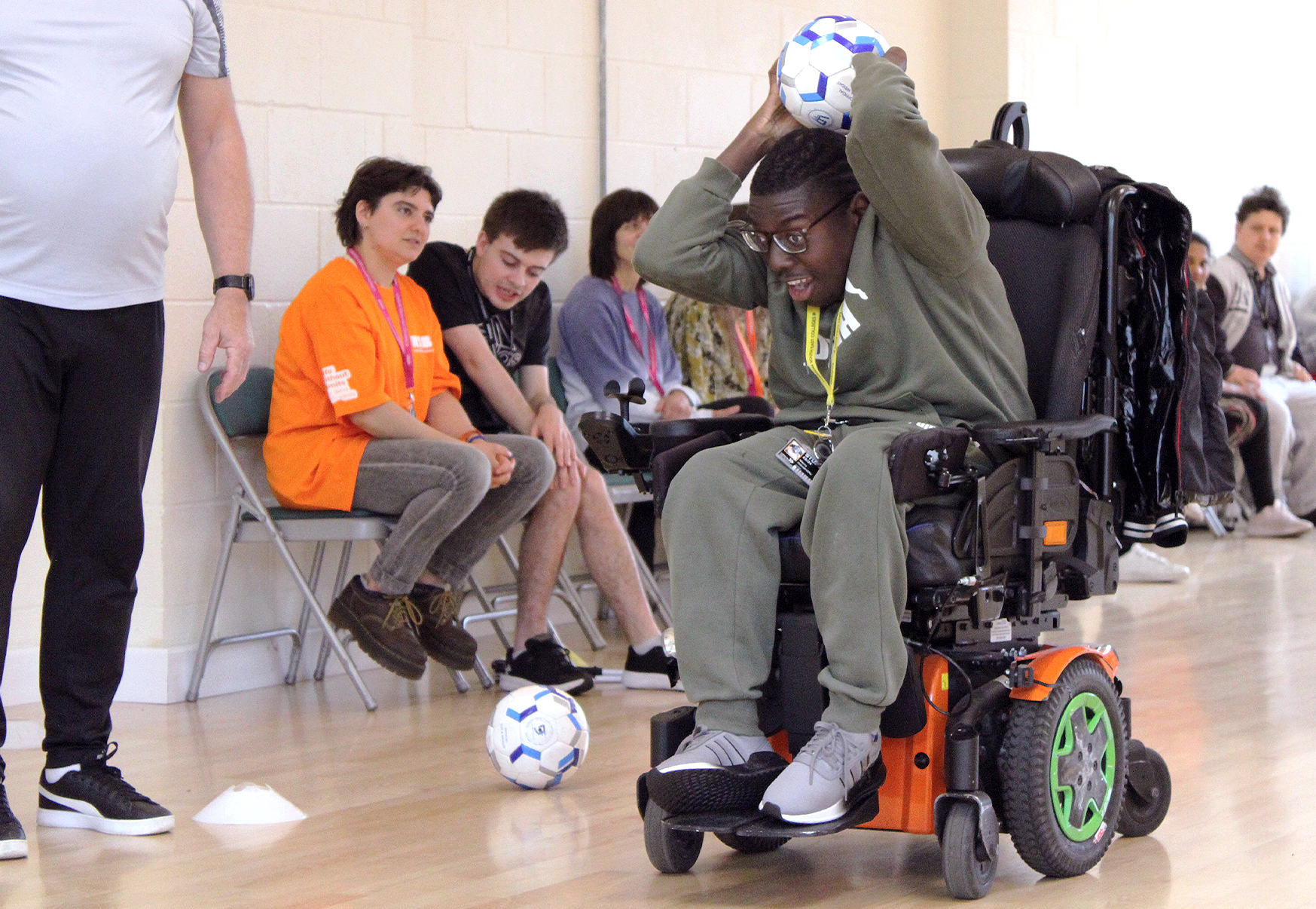 A dark-skinned young boy in a motorised-wheelchair. He is holding a ball aboe his head, preparing to throw it. His face is full of concentration.