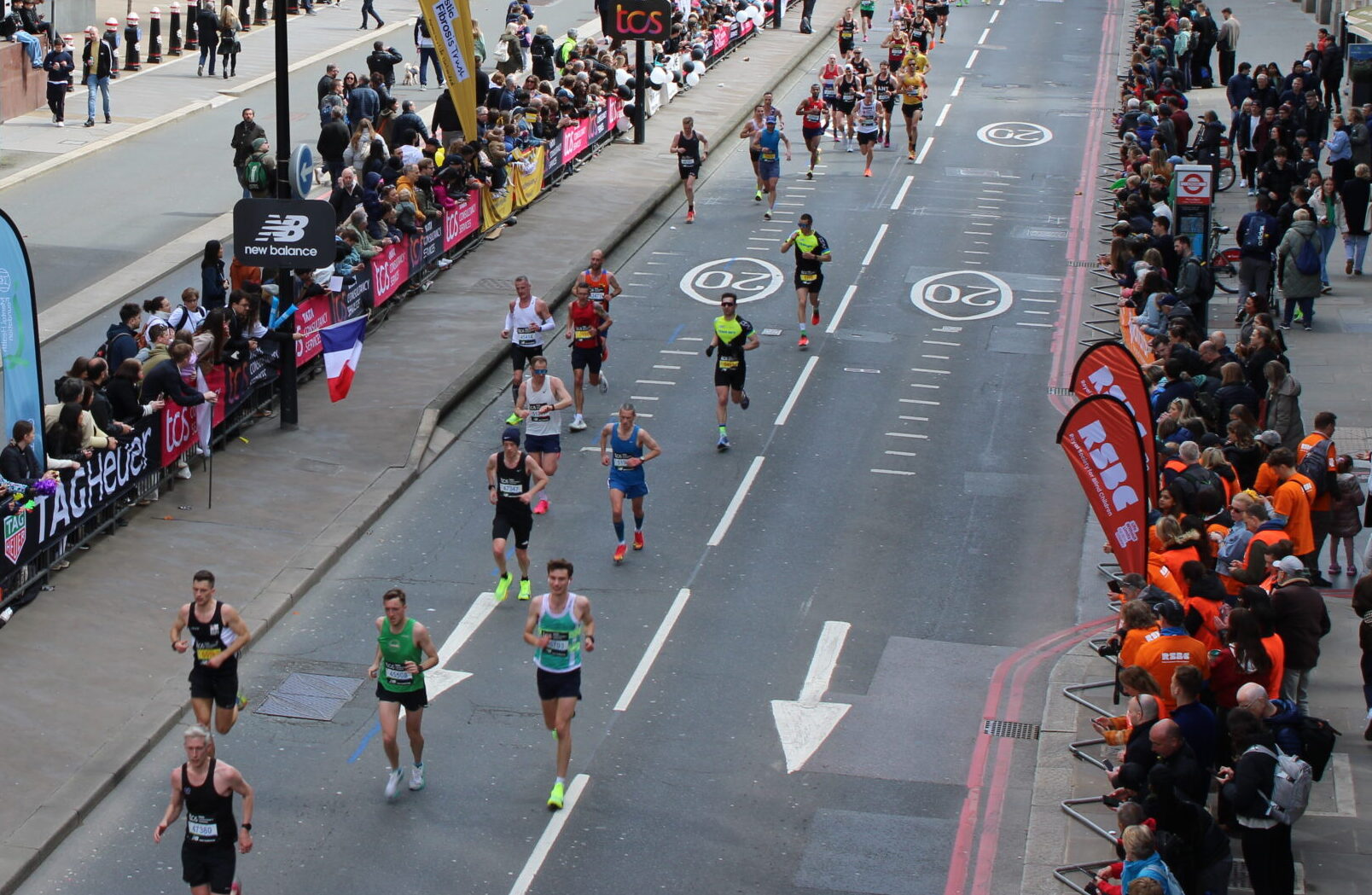 A street on the London Marathon route, shot from above. On the street is a smattering of runners with cheering supporters lining the street.