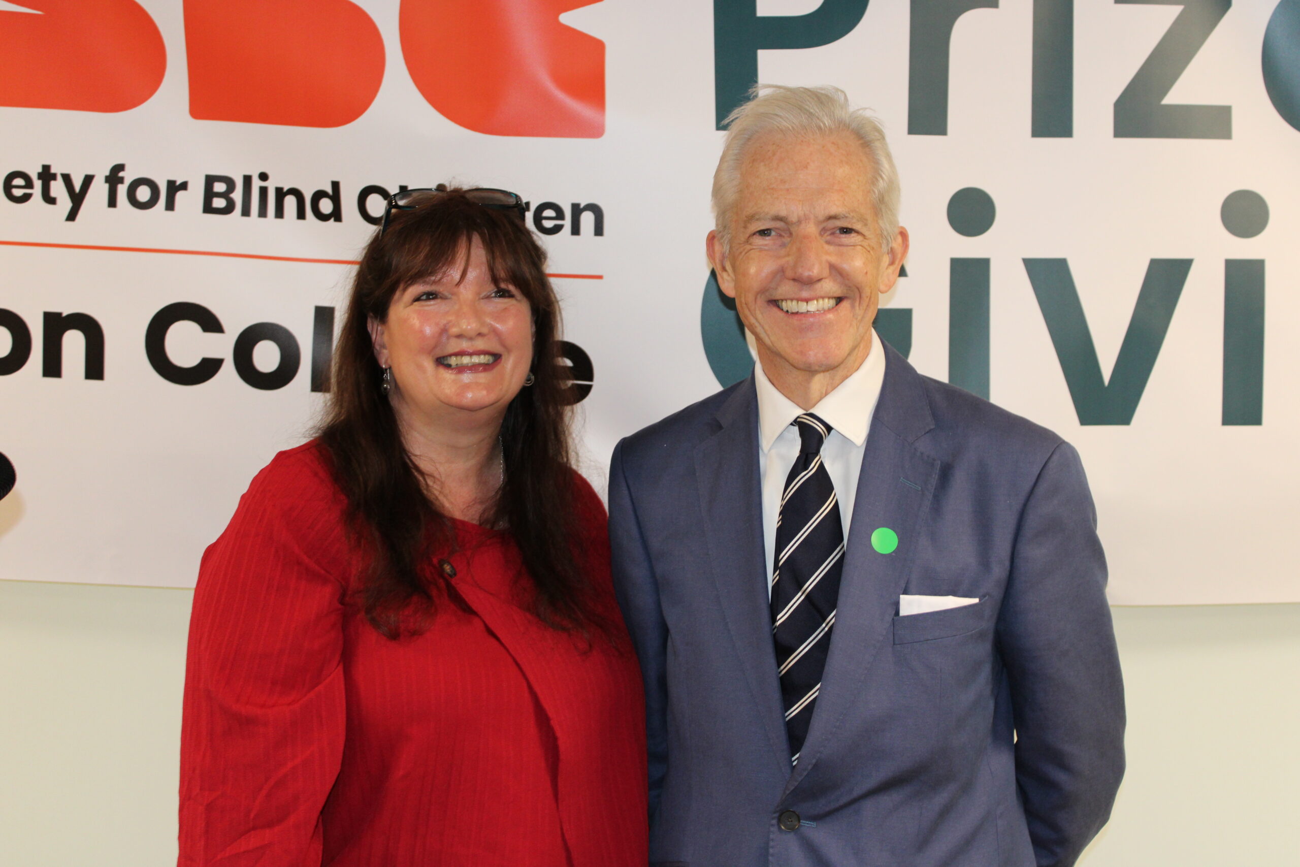 Two people standing in front of a decorative banner. On the left, a light-skinned woman with long brown hair and a fringe. On the right, a light-skinned man with short grey hair.