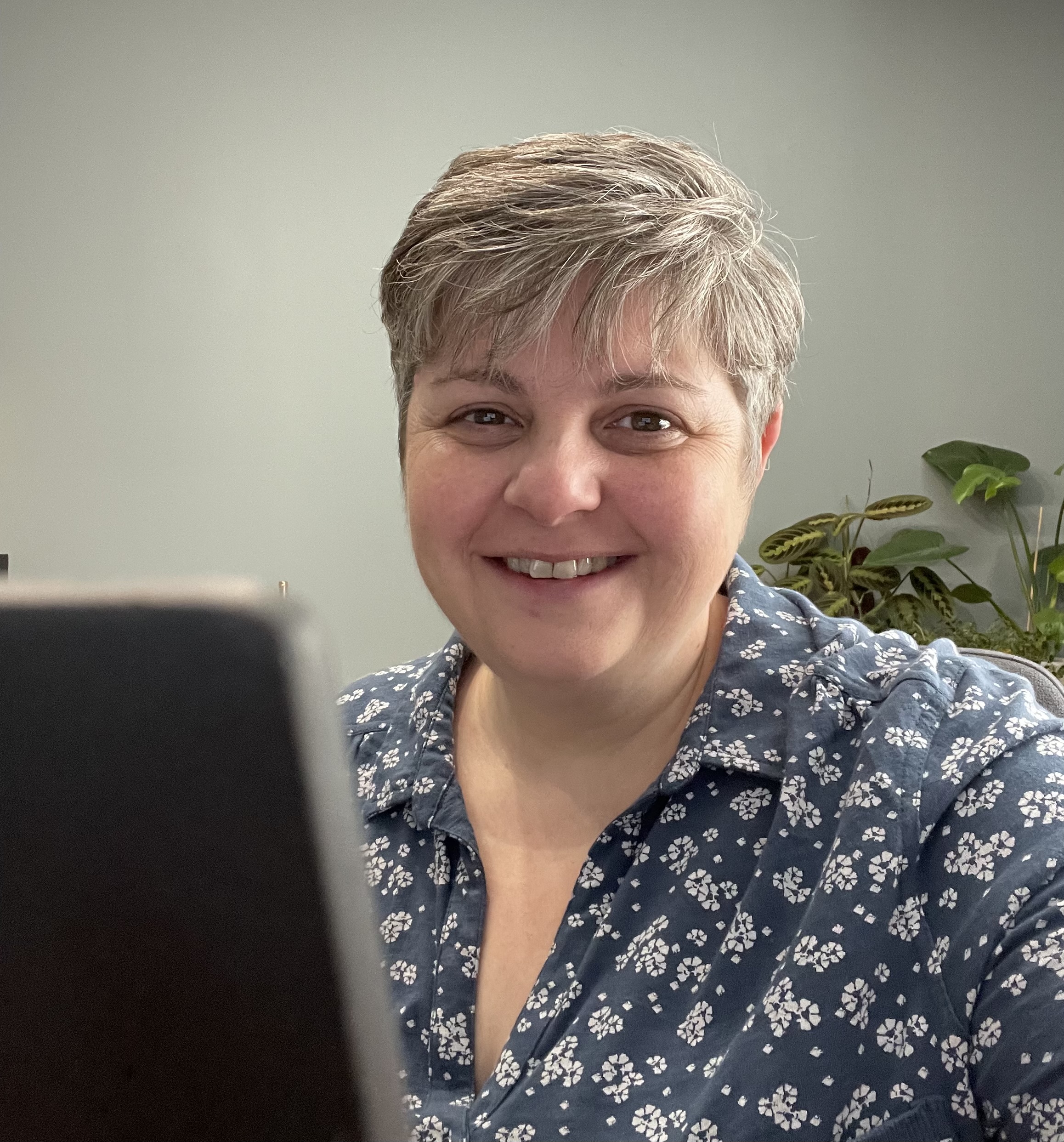 A close-up of Jeanette, a light-skinned woman with short grey hair. She is wearing a blue floral pattern shirt and is sitting in front of her laptop. Jeanette is smiling brightly at the camera.