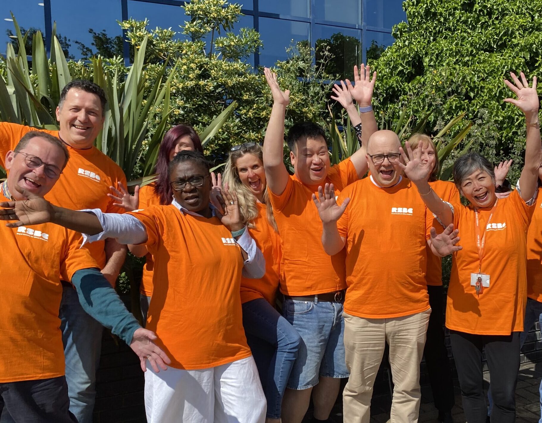 A group photo of RSBC staff members standing outside of the LWLC building. They are all wearing orange t-shirts while smiling and waving at the camera.