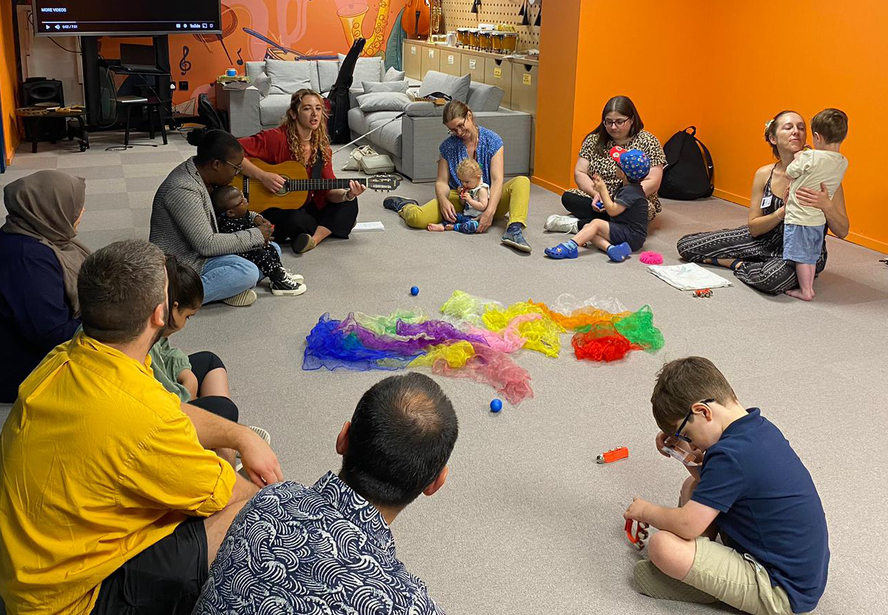 Parents and 0-7year old children seated in a circle in a large room listening to a lady playing a guitar and singing songs in a large room.