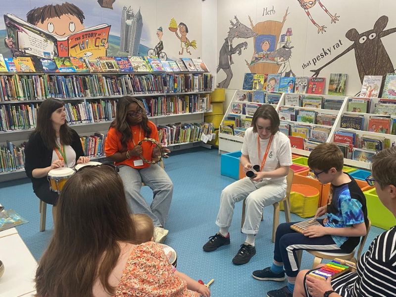 RSBC staff with parents and young children in a circle at a library. The team and the young participants are holding musical instruments.