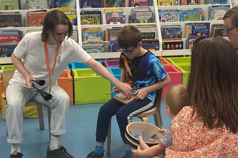 Five people (a female tutor, a young boy, a mother and a mother holding her baby) sat in a library holding and playing percussion instruments.