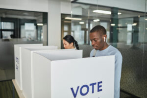 Two people at a polling station. They are casting votes in polling booths with partition walls that come up to them at chest height. Over the tops of the polling booths we can see, a medium-light skinned person with long plaited black hair in one booth and a dark-skinned person with short black hair and a mustache.