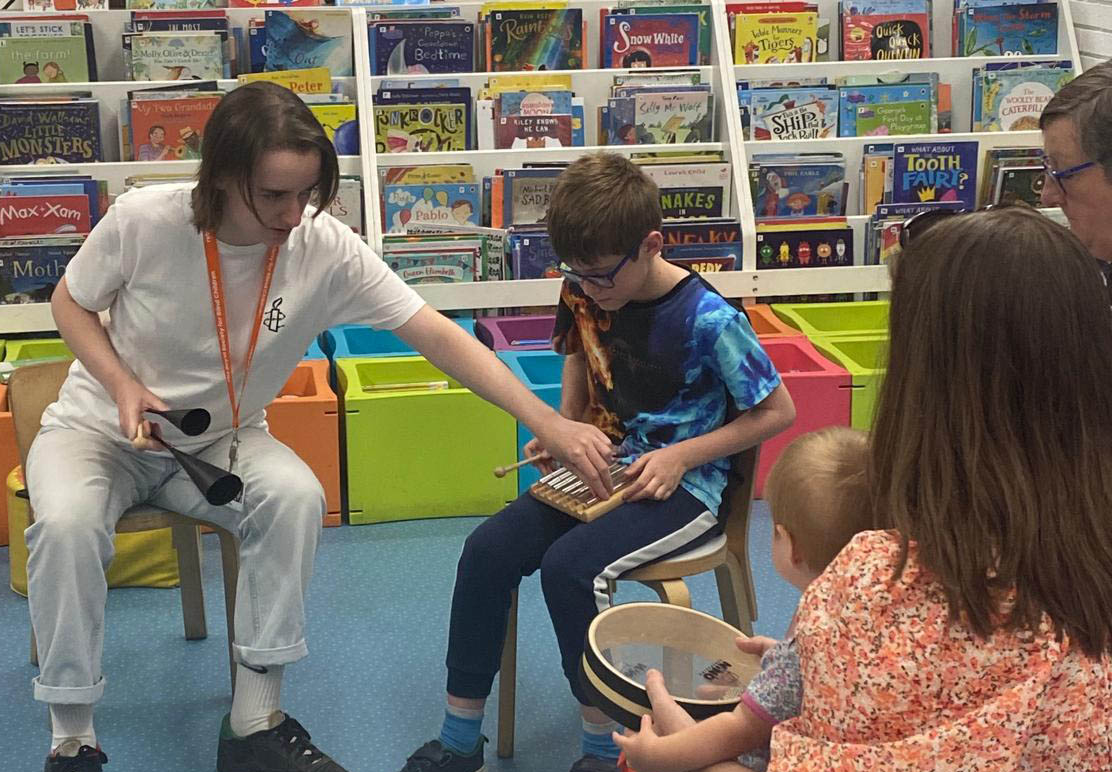 Five people (a female tutor, a young boy, a mother and a mother holding her baby) sat in a library holding and playing percussion instruments.