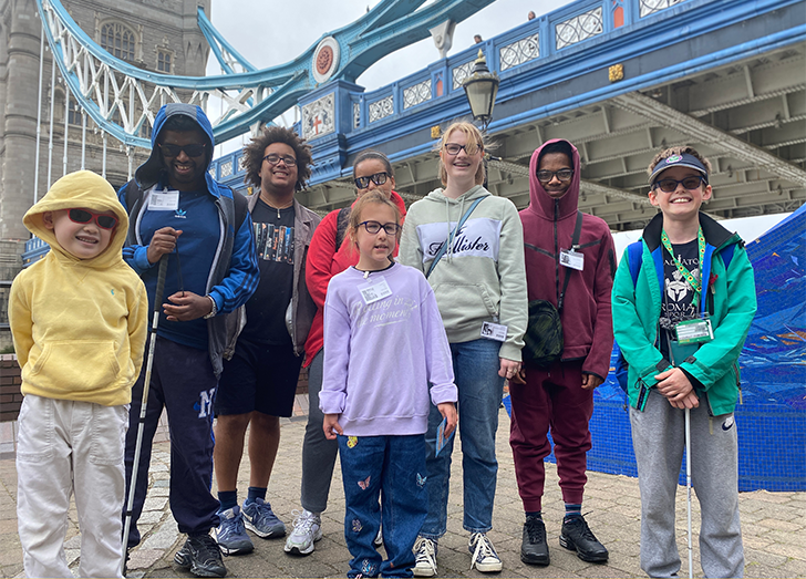 A diverse group (8) of children, young people and an adult standing in front of Tower Bridge in a group pose, smiling and looking at the camera. A boy with dark glasses and and cap, and a young person wearing a Parka coat are both holding white canes.