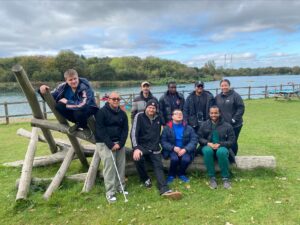 A group of young men on a residential trip with RSBC. The group are sat, huddle around an arrangement of tree logs. In the background is lush greenery and a lake.