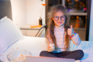 A young girl sat on a double-bed with a laptop and a big bowl of popcorn, getting ready for movie night. The girl is light-skinned with long brunette hair, wearing glasses with large circle lenses. She is smiling at the camera and posing with her thumb up.