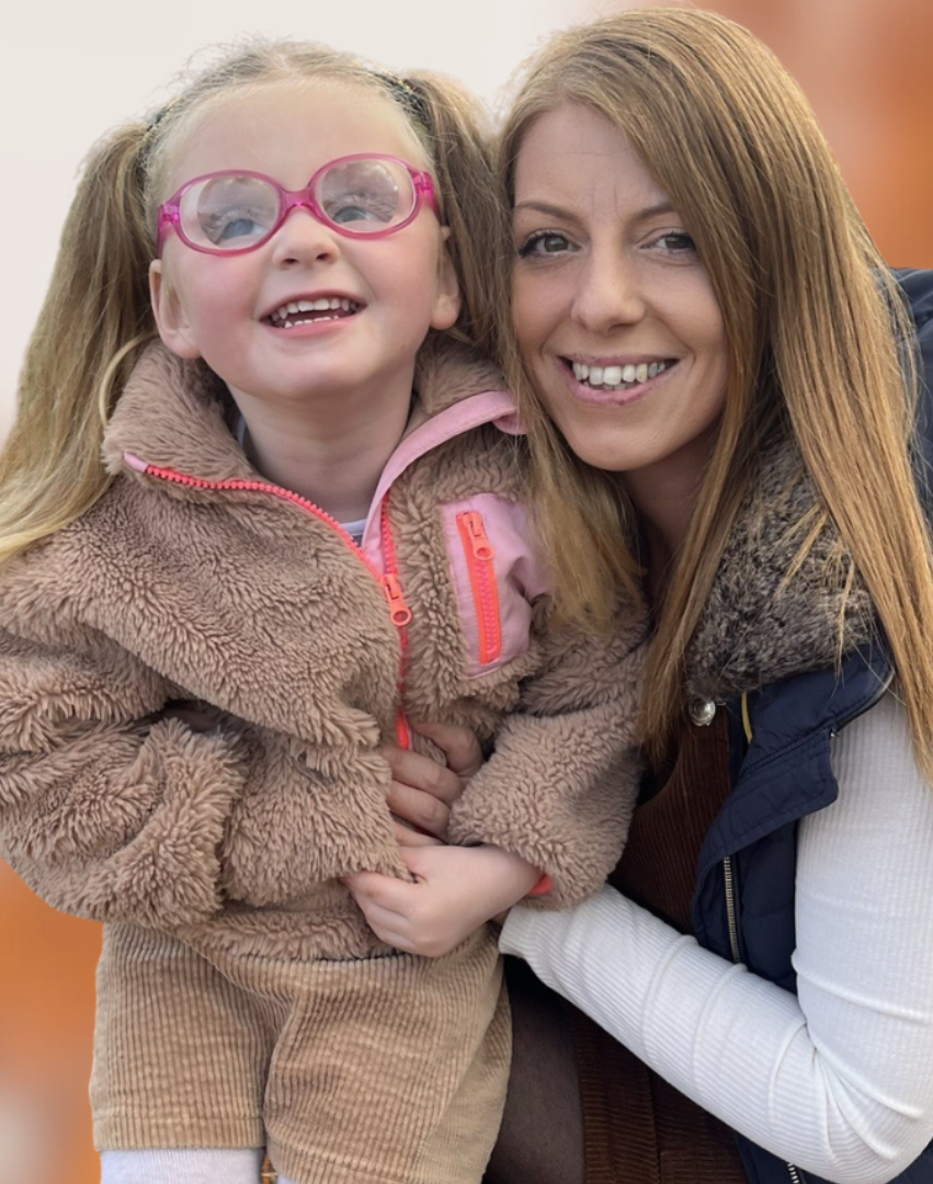 An adorable little girl being cuddled by her mum. The girl is light-skinned with long dark-blonde hair, worn in two pigtails. She has pink glasses and beige coat. Her mum is a light-sknned woman with light-brunette hair. She is wearing a black puffer-best and a white long-sleeved t-shirt. Both are smiling at the camera. Behind them is a blurred background with autumn trees.