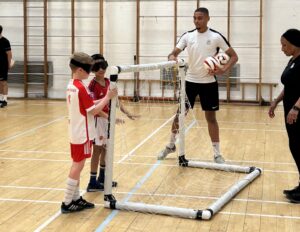 Two kids and a coach at the RSBC X Bloomsbury Football session. The two children are wearing red football kits and have black blindfolds over their eyes. Standing close to together, they are feeling out for for the goal in front. In the background, a football coach helps them navigate the space.