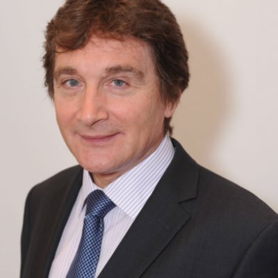 A headshot of Tom Pey, an RSBC Vice President. Tom is a light-skinned man with short chestnut brown hair. He is wearing a black blazer with a blue pin-striped shirt and dark blue tie. He is smiling warmly at the camera.