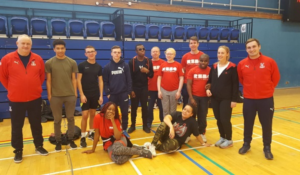 A group photo of participants and tutors at the Crystal Palace sports session. The group are standing in a sports hall huddled together, two of the young people at the front are laying on their sides, posing.