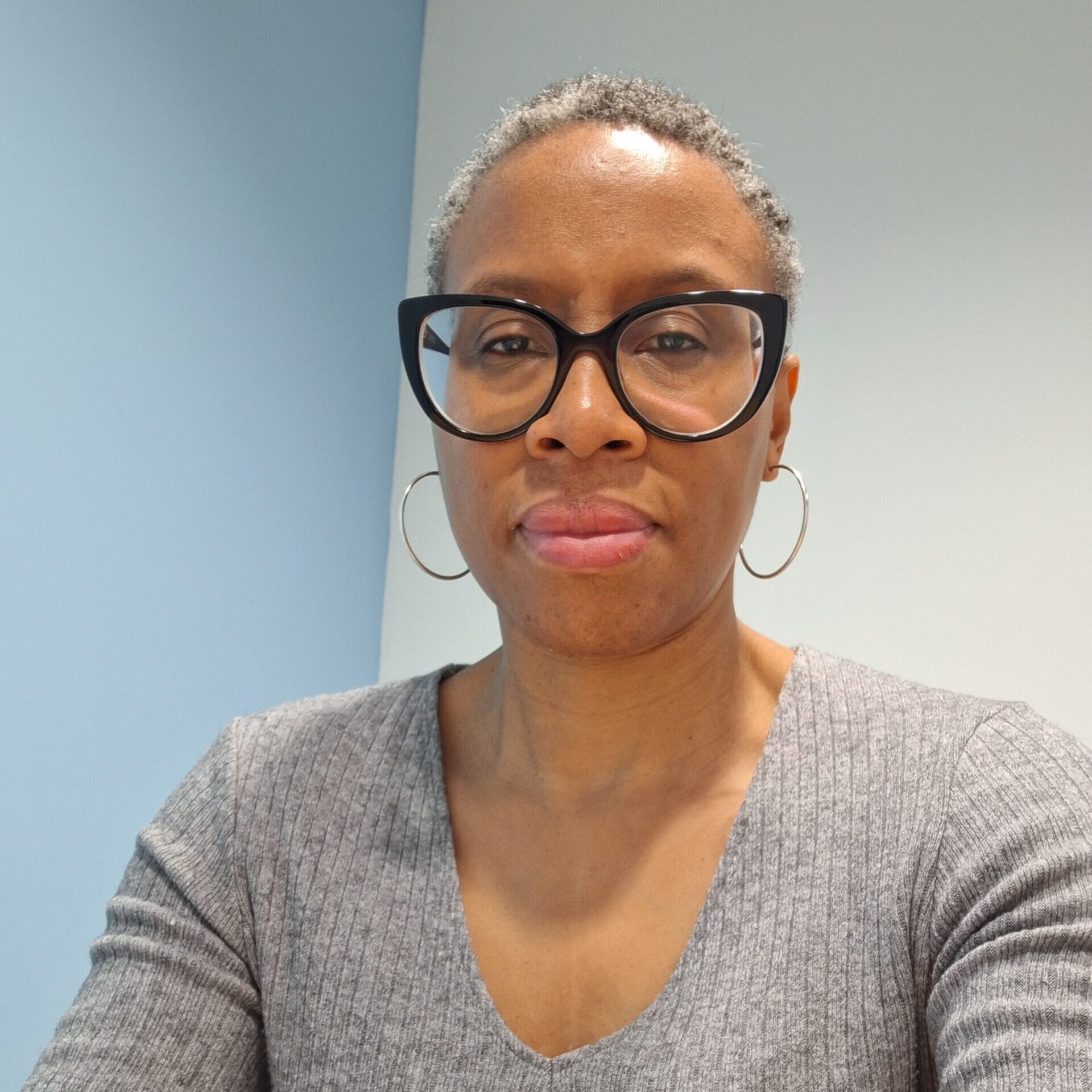 Headshot of Kathy, the PA to CEO/Office Administrator at RSBC. Kathy is a dark-skinned woman with dark grey short shaved hair. She is wearing an light-grey shirt, black glasses and silver hoop earrings. She is looking straight at the camera and smiling softly.