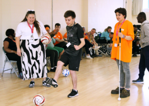 Snapshot of a Dorton College activity. A young VI light-skinned man wearing a black t-shirt, black shorts and black & white trainers is in the centre of the picture. He is having fun kicking a football. Other Dorton College students are sat in the background behind him. On the left side of the picture is a Dorton College Staff member lady smiling and holding a football under her left arm. She is wearing a white t-shirt, a black and white pattern midi-skirt and black & White Nike Dunks. On the right stands Atrix, Dorton College's Learning Support Advisor holding a cane and looking at the young man. They are wearing an oversized bright orange RSBC t-shirt, skinny jeans and brown low Dr Martins shoes.