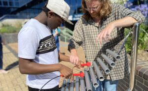 Owain from the RSBC team and a young person standing outside, exploring one of the percussive play outdoor instruments. The two people are standing either side of the instrument. The instrument is made of a series of steel grey tubes, which the young person is tapping to make sound.