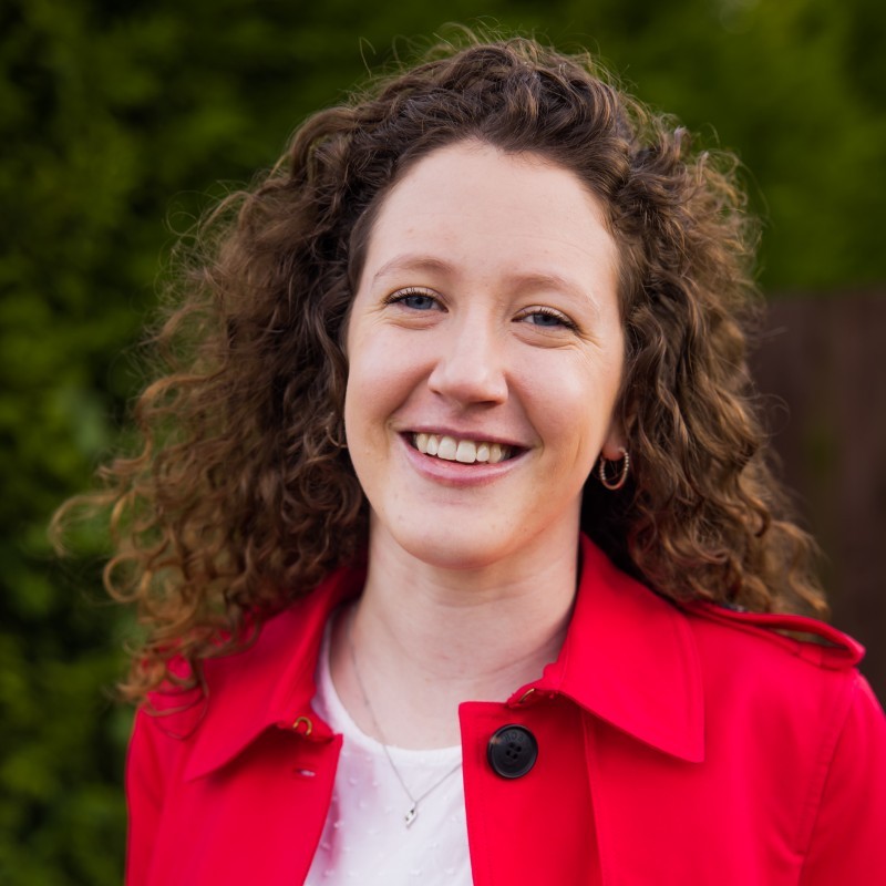 Headshot of Helen, one of the Marketing Executive & Business Partners at RSBC. Helen is a light-skinned woman with warm brown medium-length hair with voluminous curls. She is wearing a white t-shirt with a bright red coat, and is smiling at the camera.