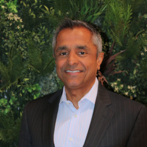 Headshot of Narayan Iyer, an RSBC Trustee. Narayan is a dark-skinned man with short dark-grey hair. He is wearing a white shirt with a black blazer. Narayan is standing in front of a wall of green foliage, smiling brightly at the camera.