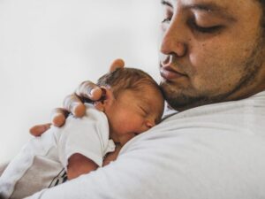 A father holding his newborn baby baby. The man is medium-light skinned with short hair and stubbed facial hair. The baby is sleeping blissful on his chest.