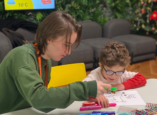 Young woman RSBC worker wearing olive green sweater helping young child with glasses colouring Christmas themed drawing.