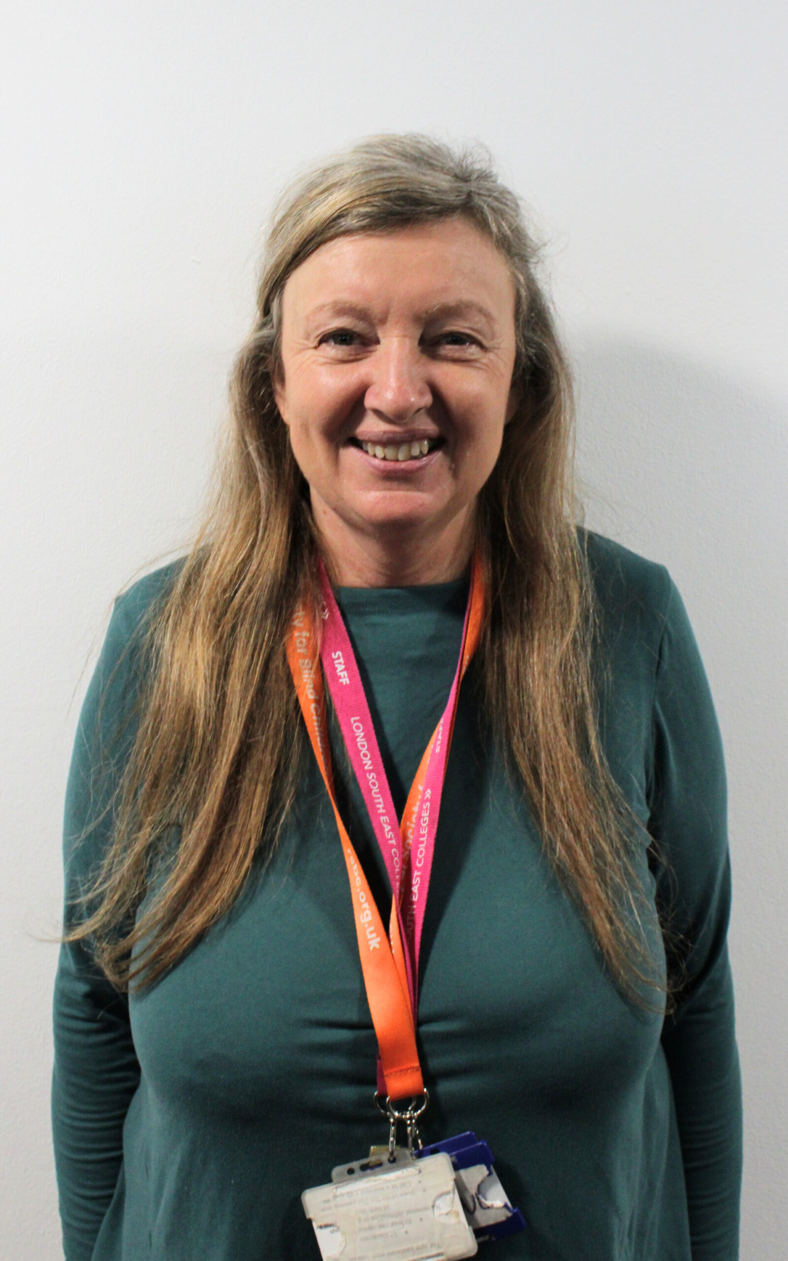 Janine, a light skinned woman with long blonde hair styled half-up half-down. She is standing in front of a white wall and is smiling while looking straight ahead. She is wearing a dark teal long-sleeved top as well as a pink and an orange lanyard.