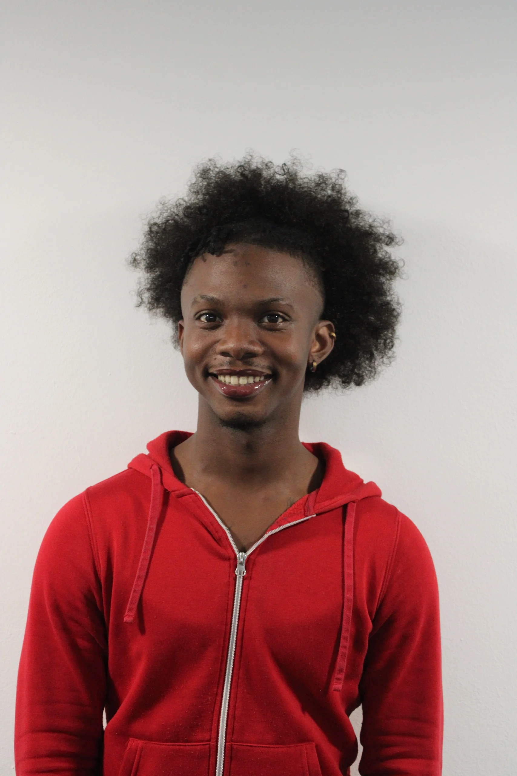 Kishawn is a dark-skin young man with coily hair styled in a ponytail. He is wearing a red hoodie zip jacket as well as two gold earrings on his left ear. He is looking at the camera with a big smille.