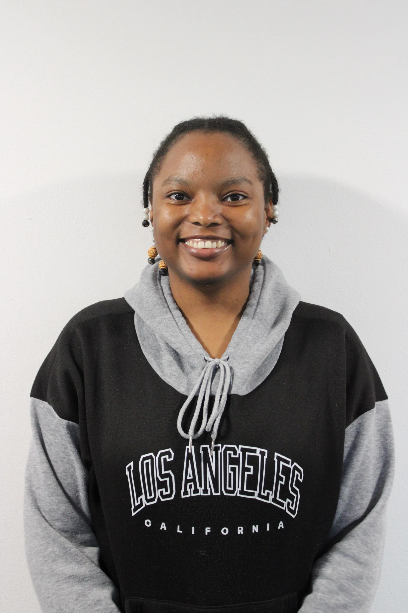 Monique is a brow skin woman. She is wearing a black and grey hoodie. Her coily hair is styled in beaded short braids. She is looking at the camera with a big smile on her face.