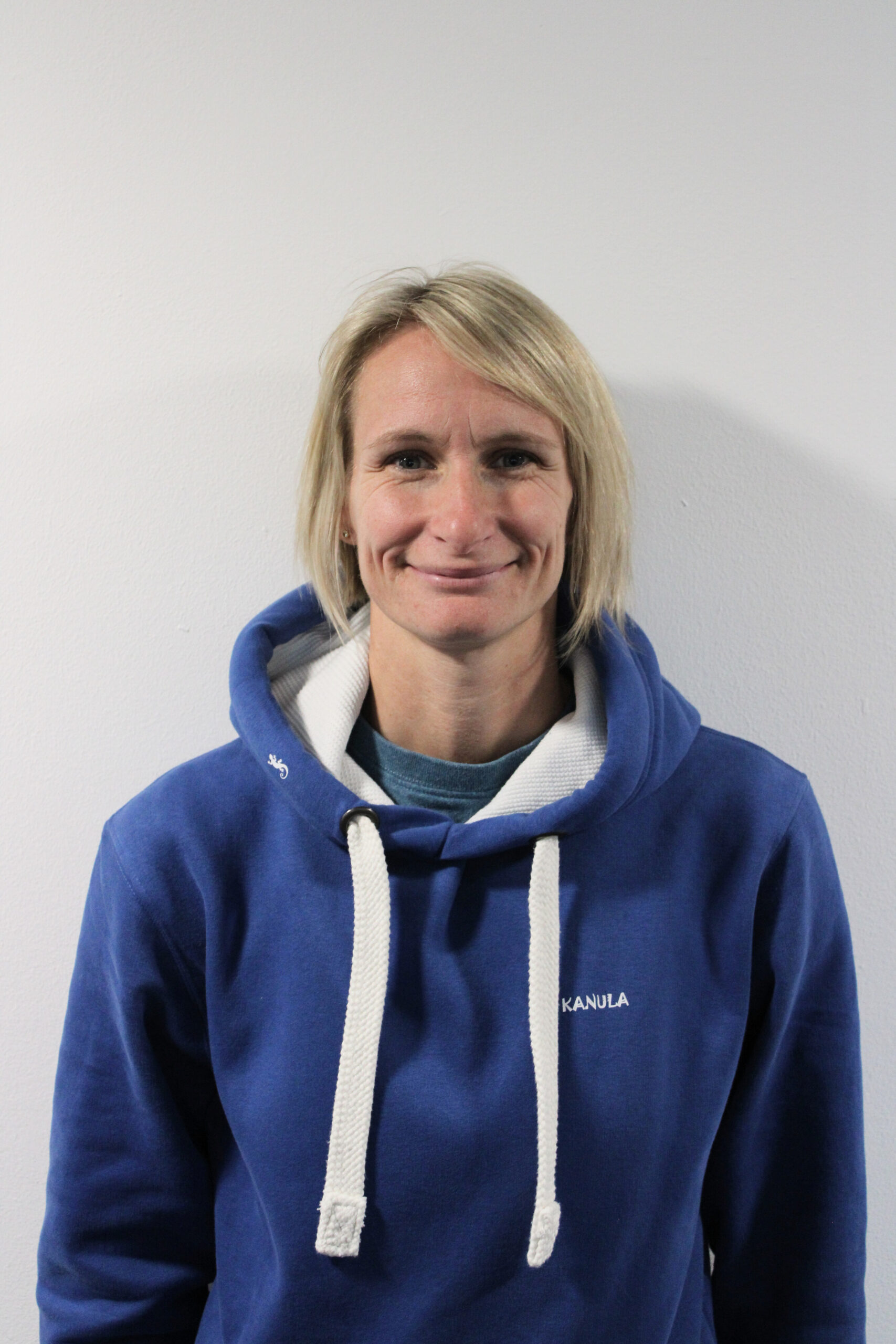 Tereza, a light-skinned woman with a shoulder length light blonde bob hairstyle. She is standing in front of a white wall and is smiling at the camera. She is wearing a blue hoodie with white strings and a hoodie with white lining.