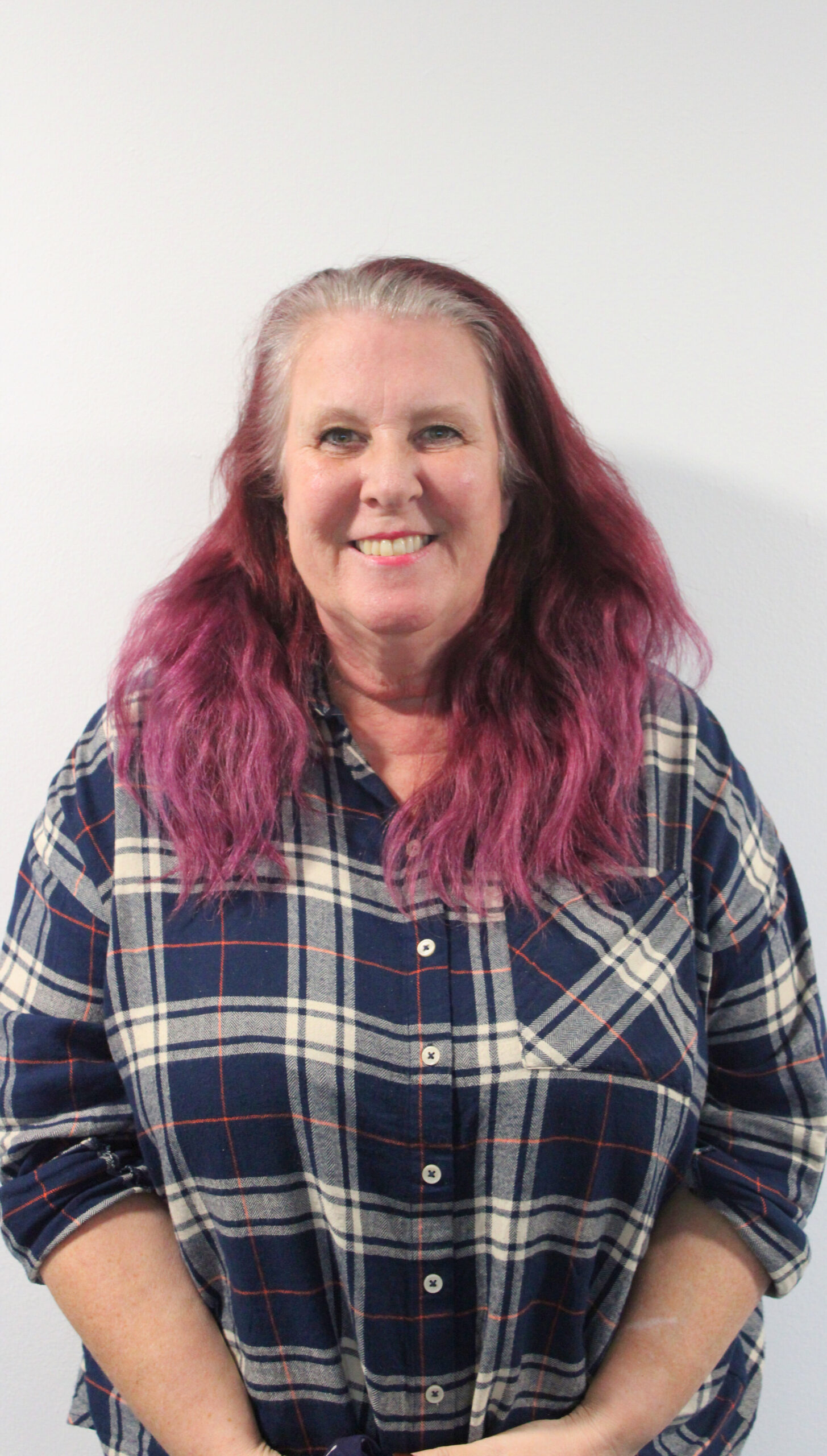 Headshot of Tricia, a Futures Tutor at RSBC Dorton College. Tricia is a light-skinned woman with purple brown medium-length hair. She is standing in front of a white wall and is smiling warmly at the camera. She is wearing a blue shirt with white tartan motif.