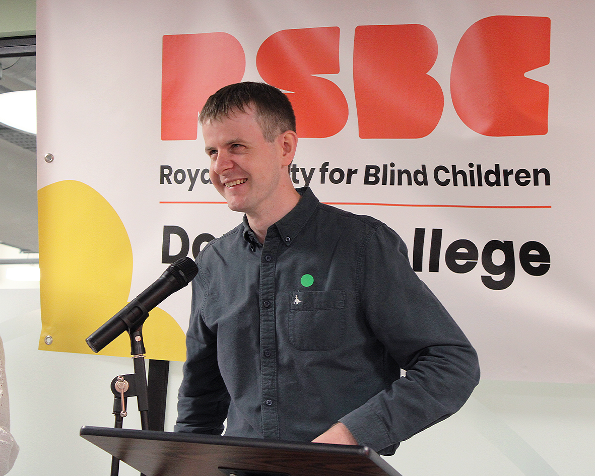 Callum, a light skinned man with dark hair, wearing a grey long sleeved shirt with black buttons. He is standing behind a podium lecture desk, with a microphone attached to the top of the desk. Callum is standing in front of a white banner with RSBC written in large orange text, and Royal Society for Blind Children written in small text underneath, there is a thin orange line underneath this text. Underneath the orange line Dorton College is written in black medium sized text. In the bottom left hand corner there is a yellow shape similar to two halves of a flower petal.