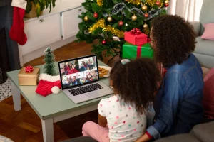 Mother and daughter with curly textured hair making laptop video call with friends and family. The laptop is on a green table. There is a green Christmas tree in the background and a fire place with a red and white stocking on it.