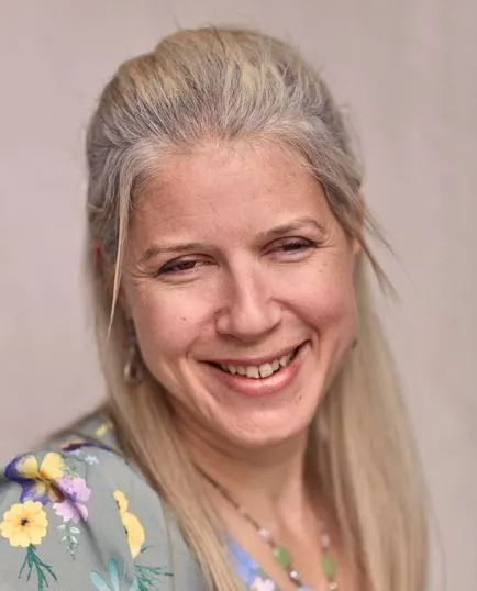 A side portrait photo of a smiling light-skinned women with light blonde hair. She is wearing a light green floral dress with yellow and pink flowers on it.