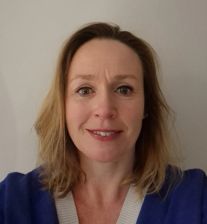 A mid-shot of a light skinned women with dark blonde hair looking directly at the camera. She is wearing a blue v-neck jumper with two white panels on the inner v.