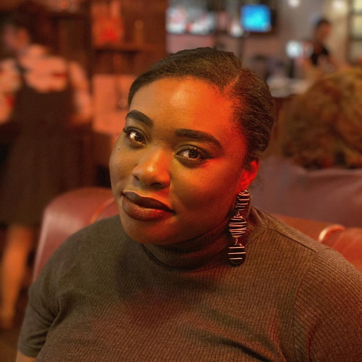 Headshot of Sharon, a dark-skinned woman with dark brown hair tied back in a ponytail. Sharon is wearing a black turtle-neck t-shirt, geometric earrings and has burgundy lipstick. She is smiling softly at the camera.