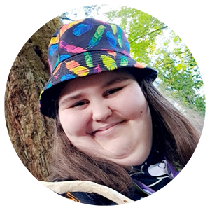 Molly is a light-skin young woman with dark brown eyes and shoulder length light brown hair. She is wearing ablack bucket hat with a colourful feathers pattern and black jumper. She is looking directly at the camera and smiling. She is standing in a sunny forest.